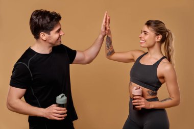 İki fitness meraklısı yoğun antrenmanlarını bir beşlikle kutluyor, her biri protein içecekleri tutuyor, sağlıklı bir yaşam tarzına olan bağlılıklarını sergiliyor ve coşkuyla diğerlerine ilham veriyor.
