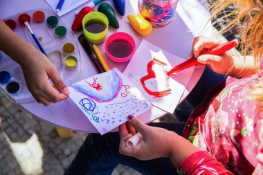 İki çocuk, açık hava masasında çeşitli malzemeler kullanarak renkli sanat eserleri çizerek yaratıcılıklarını ifade etmekle meşguller. Ortam neşeli ve davetkar..