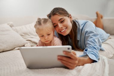 Sevgi dolu bir anne ve tapılası bebeği, yumuşak yataklarında rahat bir şekilde, tablet üzerinde büyüleyici ve eğlenceli bir şeyler izleyerek, bir an paylaşıyorlar.