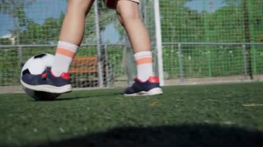 Bu canlı açık hava sahnesinde, turuncu şort ve beyaz çoraplı küçük bir çocuk futbol oynarken görülüyor yemyeşil çimenler ve bir futbol kalesinin günlük düzenlenmesi eğlenceli ve enerjik bir spor gününün müjdecisi.