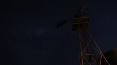 Geniş yıldızlı bir gece göğüne karşı geleneksel bir yel değirmeni siluetinin büyüleyici zamanlaması. Sahne, yıldızların hareketlerini ve kırsal bölgenin barışçıl atmosferini yakalıyor..