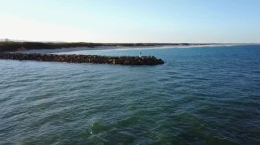 Küçük bir deniz fenerine giden büyük kayalarla kaplı bir dalgakıranın yer aldığı Avustralya kıyı şeridinin havadan görünüşü. Huzurlu mavi okyanus yapıyı sarar..