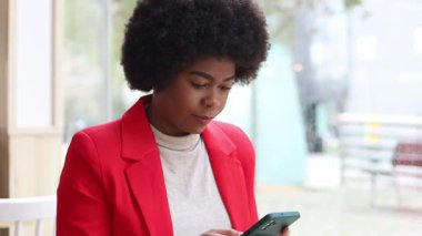 Afro saçlı Afrikalı Amerikalı bir iş kadını cep telefonuyla modern bir kafe ortamında buluşuyor, şık kırmızı bir ceket giyiyor..