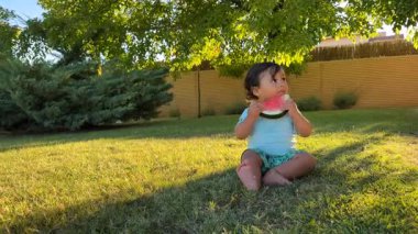 Küçük bir erkek bebek çimlerin üzerinde oturmuş, büyük bir dilim sulu karpuzu ısırıyor. Güneşli bir arka bahçede yaz ziyafetinin tadını çıkarırken ifadesi çok keyifli..