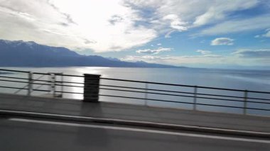 Lavaux, İsviçre 'de, Cenevre Gölü manzaralı ve uzak dağları olan bir yol. Güneş ışığı suya yansıyor ve bulutlar geniş ve huzurlu bir manzara yaratıyor.