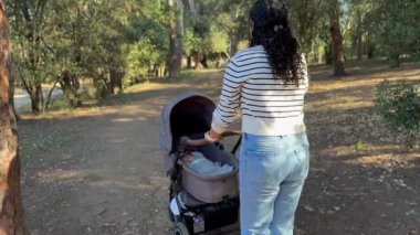 Bebek arabasını iterken manzaralı bir parkta yürüyen bir kadının arka görüntüsü. Mekan uzun ağaçlar ve toprak bir patikayla huzur dolu. Bu da dışarıda huzurlu bir gün geçirdiğimizi gösteriyor..