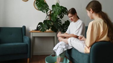 Bir anne, ayaklarını nazikçe temizleyici bir ürünle ovalıyor. İkisi de ayaklarını portakal dilimleriyle süslenmiş bir kovaya sokuyor. Bir spa günü boyunca rahatlatıcı ve samimi bir anı paylaşıyorlar..