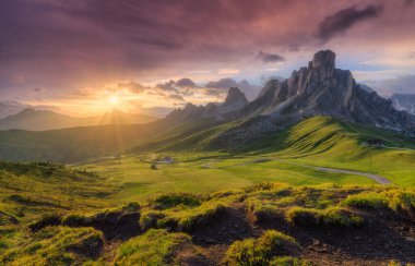 Bereketli yeşil vadi, yazın renkli günbatımında güzel alp dağları. Passo Giau, Dolomitler, İtalya. Yeşil çayır, yol, yüksek kayalar ve bulutlu pembe gökyüzü altın gün batımında