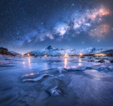 Yıldızlı kış gecesinde fiyort ve karlı dağların üzerindeki Samanyolu kemeri. Lofoten Adaları, Norveç. Yıldızlı gökyüzü, buzlu donmuş deniz yansıması, samanyolu, şehir ışıkları, karlı kayalar. Boşluk