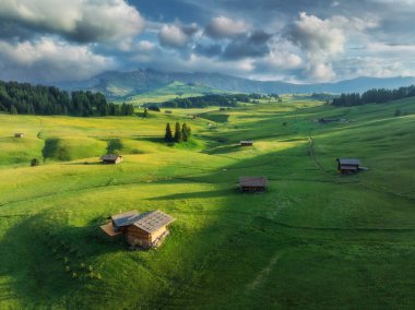Avrupa 'nın en büyük yüksek irtifa dağlık çayırının havadan görünüşü, Alpe di Siusi, gün batımında yemyeşil bir arazide yuva yapmış ahşap kulübeler. Dağlardaki evlerin en üst görüntüsü. İtalya 'da yaz mevsimi