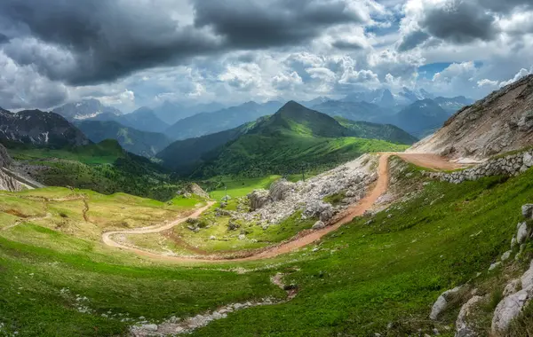 Parlak yeşil bir vadide kıvrımlı dağ yolu, yazları zıt ışık ve gölgelerle dolu dramatik bir gökyüzünün altındaki dağlık tepeler. Panoramik renkli manzara. Dolomitler, İtalya. Doğa