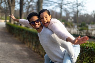 Genç adam parkta kız arkadaşını sırtında taşıyor, kollarını açmış ve gülüyor.