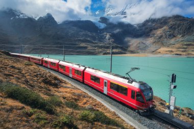 Kızıl tren sonbaharda mavi gökyüzü olan güneşli bir günde İsviçre Alp Dağları 'nda, göl kenarında demiryolu boyunca ilerliyor. Manzara. Bernina Ekspres treni, turuncu çimenler, sonbaharda gün batımında karlı kayalar. Seyahat