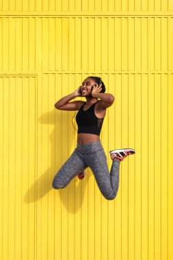 Sarı duvara karşı atlayan ve güneşli bir günde eğitim sırasında kulaklıkla müzik dinleyen aktif giysili neşeli Afrikalı Amerikalı genç bir kadın.