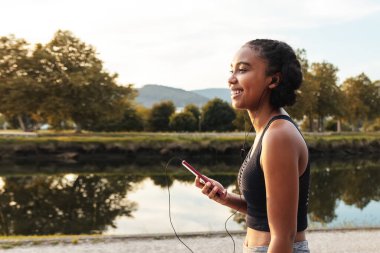 Genç, düşünceli, neşeli, Afro-Amerikan bayan sporcunun yan görünüşü. Kulaklıkla müzik dinlerken, cep telefonuyla nehrin kıyısında durup, gözlerini kaçırması.