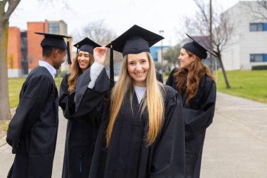 Mezuniyet elbisesi giyen, üniversite kampüsündeki mezuniyet günlerini kutlayan neşeli çok ırklı öğrencilerden oluşan bir grup.