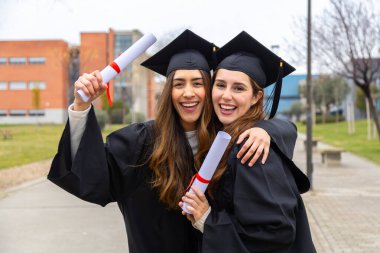 Mezuniyet elbisesi ve kep giyen iki kız öğrenci mezuniyet gününde mutlu bir şekilde diplomalarını tutuyor ve birbirlerine sarılıyorlar.