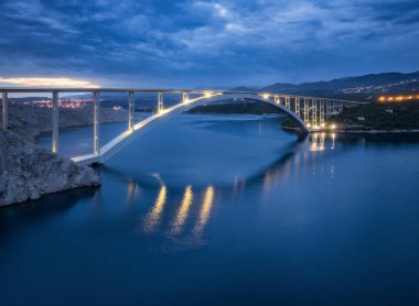 Geceleri mavi deniz ve köprü manzarası. Günbatımında aydınlık, ada, dağlar, su ve bulutlu gökyüzü olan modern Krk köprüsü. Yol manzarası, şehir ışıkları, alacakaranlıkta kayalar. Mimari