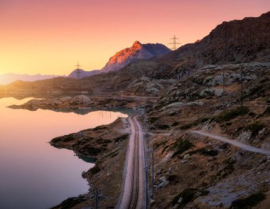 Railway station near alpine mountain Bianco lake, rocks and colorful pink sky at sunset in autumn. Bernina Express Railroad, Switzerland. Mountain valley in Alps, reflection in water in fall at dusk