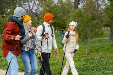 Dört arkadaş kış boyunca parkta yürüyüş yapıyor, yürüyüş direkleri ve sırt çantaları kullanıyor, konuşuyor ve gülümsüyor.