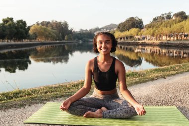 Genç içerikli Afro-Amerikan kadın bacak bacak üstüne atmış halde nehir kenarındaki minderde yoga yapıyor ve gözlerini kaçırıyordu.