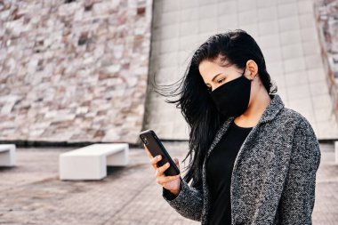 Şehir sokaklarında salgın sırasında kaldırımda dikilirken akıllı telefon kullanan ve gözlerini kaçıran maskeli etnik bir kadın.