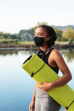 Spor giyim ve maske takan Afro-Amerikalı genç bir bayanın sabah antrenmanından önce havuz kenarında ellerinde hasırla yürümesi.