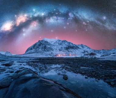 Gece yıldızlı gökyüzü Samanyolu kemeriyle, suda taşlar. Samanyolu ve yıldızlı kış manzarası aydınlık, fiyort, sahil, deniz ve karlı dağları olan rorbu evleri. Lofoten Adaları, Norveç. Boşluk