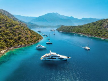 Aerial view of beautiful yachts and boats on the sea at sunset in summer. Gemiler Island in Turkey. Top view of luxury yachts, sailboats, clear blue water, beach, mountain and green forest. Travel