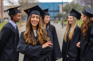 Mezuniyet töreninden sonra meslektaşlarıyla dışarıda dururken kollarını kavuşturup gülümseyen neşeli lisansüstü öğrencisinin portresi.
