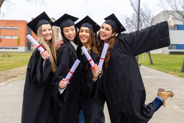 Diplomaları olan, üniversitenin dışında mezuniyetlerini kutlayan, akademik elbiseler içinde dört neşeli çoklu etnik kadın mezun.