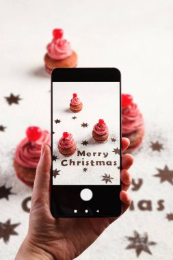 Yukarıdan gelen akıllı telefonlu isimsiz kişi renkli çöreklerin fotoğrafını çekiyor. Üzerinde krema ve kirazlı böğürtlenler ve Noel süslemeleri var.