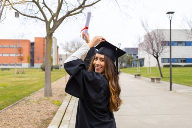 Gecelik ve havan topu giyen genç mezun, canlı bir üniversite kampüsünde diplomasını sevinçle yükselterek, başarısını kutluyor.