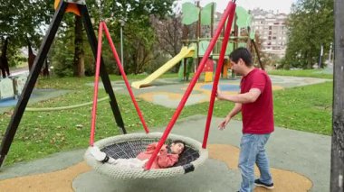 Serebral felçli reşit olmayan bir çocuk oyun parkında salıncakta sallanırken babası yavaşça salıncakta sallanırken rahat görünüyor. Çevreleri yeşillik ve oyun sahası ekipmanlarıyla çevrili olup, destekleyici ve eğlenceli bir çevreye dikkat çekerler..