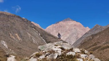 Andes Dağları 'nın engebeli arazisine tepeden bakan, kayalık bir çıkıntının üzerinde berrak mavi bir gökyüzünün altında, tanınamayan yalnız bir adamın arka görünüşü..