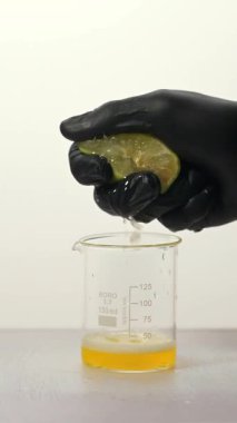 Siyah eldivenli biri limondan taze meyve suyunu şişeye sıkıyor. Ölçümlerle işaretlenmiş deney şişesi, canlı sarı bir sıvı içeriyor..