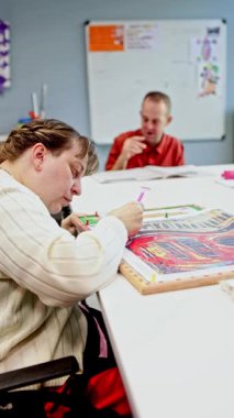 Zihinsel engelli bir kadın elmas tablosuna odaklanırken, arka plandaki bir erkek başka bir aktiviteye katılıyor, yaratıcılık ve kapsamı bir günlük merkezde ön plana çıkarıyor.