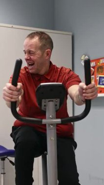 Bir gün merkezindeki terapi seansı sırasında sabit bisiklet üzerinde zihinsel engelli egzersizleri yapan bir adam, destekleyici bir ortamda sağlık, dayanıklılık ve kişisel motivasyonu teşvik eder.