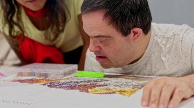Zihinsel engelli bir adam, bir bakıcı tarafından yönetilen, destekleyici ve kapsayıcı bir çevrede yaratıcılığı, odaklanmayı ve iyi motor becerileri teşvik eden bir elmas resim sanatına konsantre olur..