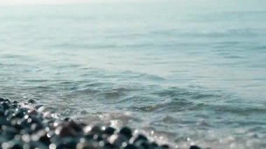 Gürcistan 'ın doğal hareketini ve ışıltısını gösteren, güneşli çakıl taşlı bir plajın üzerinden akan yumuşak dalgaları gösteren gerçek zamanlı bir video.