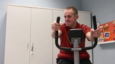 Zihinsel engelli bir adam spor salonunda egzersiz bisikletine biniyor. Formunu, motor becerilerini ve kişisel refahını geliştirmek için fiziksel aktivitelere katılıyor..