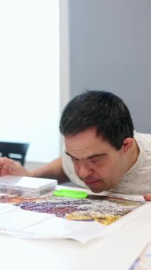 Zihinsel engelli bir adam, kapalı bir etkinlik sırasında, dikkatleri, yaratıcılığı ve kişisel başarıyı güvenli bir alanda teşvik eden ayrıntılı bir elmas sanat eseri üzerinde dikkatle çalışır.