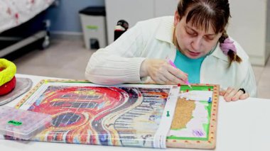 Zihinsel engelli bir kadın, sanat terapisi yoluyla yaratıcılık, sabır ve hassas motor koordinasyon geliştirmek amacıyla, bir merkezde detaylı bir elmas resim projesinde çalışıyor.