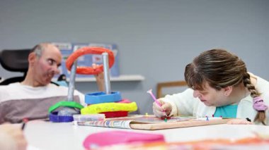 Zihinsel engelli yetişkinler bir gündüz merkezinde yaratıcı bir etkinlik oturumuna katılıyorlar, renkli sanatlara odaklanıyorlar ve terapi ve eğlence desteği olarak resim yapıyorlar..