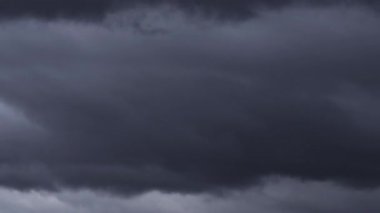 Gri gökyüzünde yoğun, karanlık fırtına bulutlarının dramatik ve hızlı bir şekilde birikmesini gösteren bir zaman kaydıyla yaklaşan şiddetli hava olaylarını sembolize ediyoruz..
