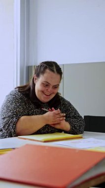 Düşük sendromlu neşeli bir kadın eğitim faaliyetlerine katılıyor, elinde kalem tutuyor ve renkli klasörlerle bir masada oturuyor, aktif öğrenme ve destek veren bir ortamda yer alıyor..