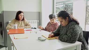 Bir bakım görevlisi, zihinsel engelli iki vatandaşı dikkatle görevlerine odaklanarak çizim faaliyetlerine katılırken denetlemektedir..