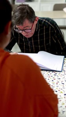 Tanımlanamayan bir bakım işçisinin zihinsel engelli bir asistana okumada yardım etmesi, zihinsel sağlık merkezindeki etkileşimlerini ve desteklerini vurgulaması. Sahne, terapi çalışmalarındaki özeni ve bağlılığı yansıtıyor..