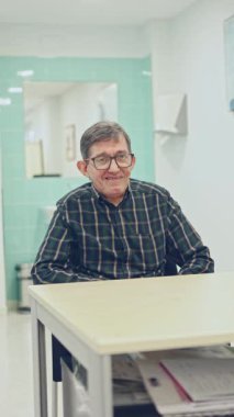 Gözlüklü ve ekose gömlekli mutlu bir yaşlı adam parlak, temiz bir ruh sağlığı konutunda bir masaya oturur, dingin bir ortamda izleyiciye sıcak bir şekilde gülümser..