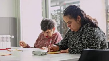 Zihinsel engelli iki kadın vatandaş, zihinsel gelişim faaliyetlerine katkı ve konsantrasyon göstererek, zihinsel sağlık merkezindeki bir masada çizim ve renklendirme faaliyetlerine odaklanmıştır.